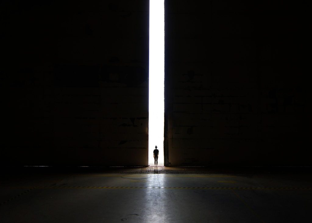 Silhouette einer Person vor einem schmalen Lichtspalt in dunklem Raum als Symbol für Hoffnung und den Weg aus der Angst